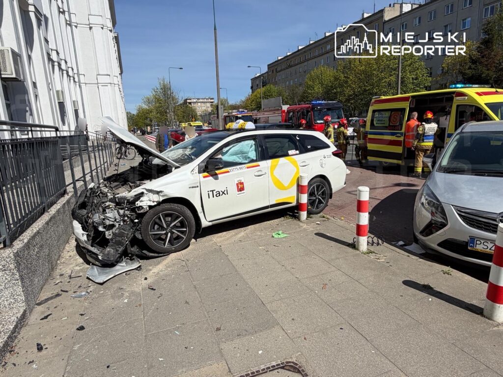 Wybuchnięty samochód taxi z uszkodzoną przednią częścią stoi przy chodniku, w tle widać strażaków i karetkę.