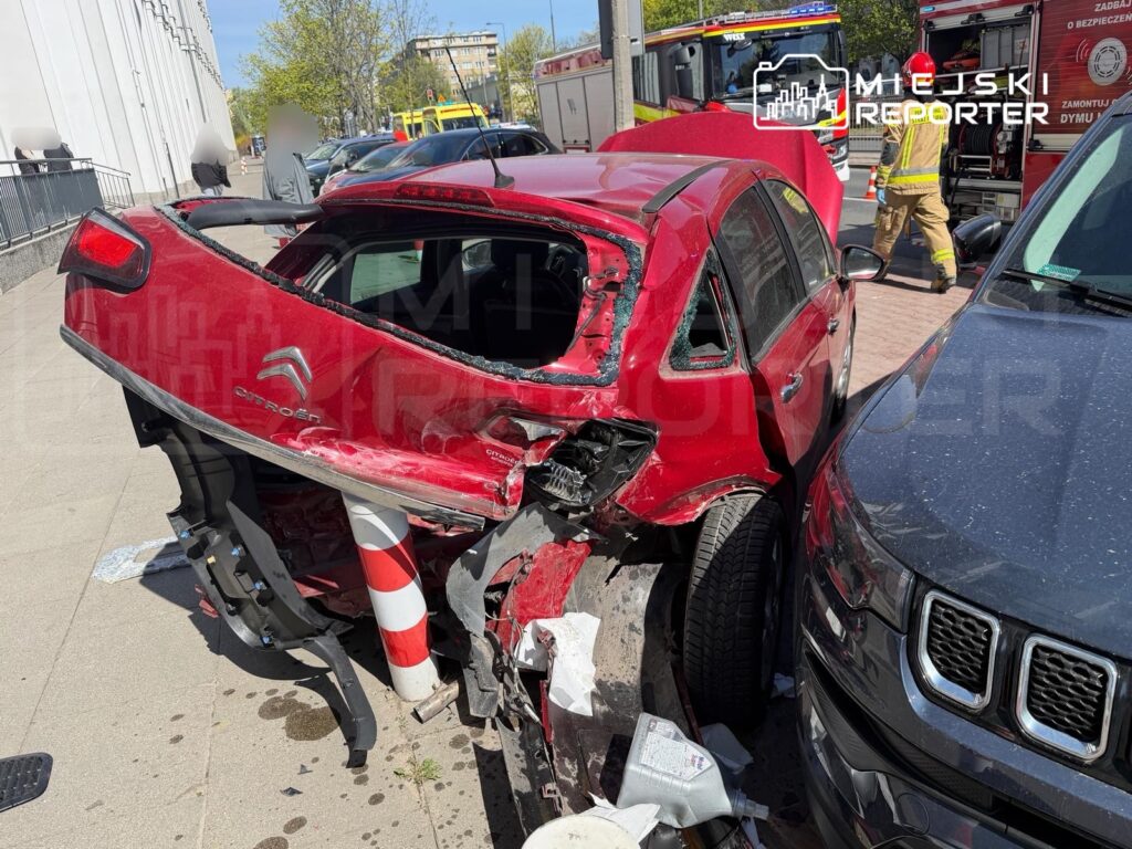 Czerwony samochód osobowy z uszkodzoną tylną częścią stoi przy krawężniku obok drugiego pojazdu, w tle strażacy w akcji.