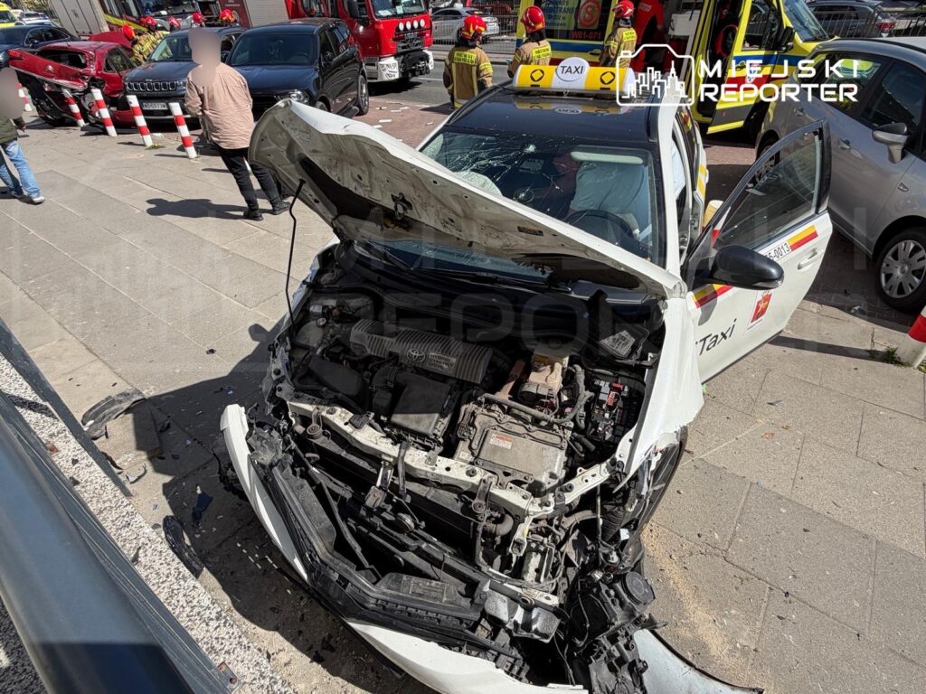 Zniszczony taksówka z otwartą maską stoi na chodniku, wokół widać inne uszkodzone pojazdy oraz osoby obserwujące zdarzenie.