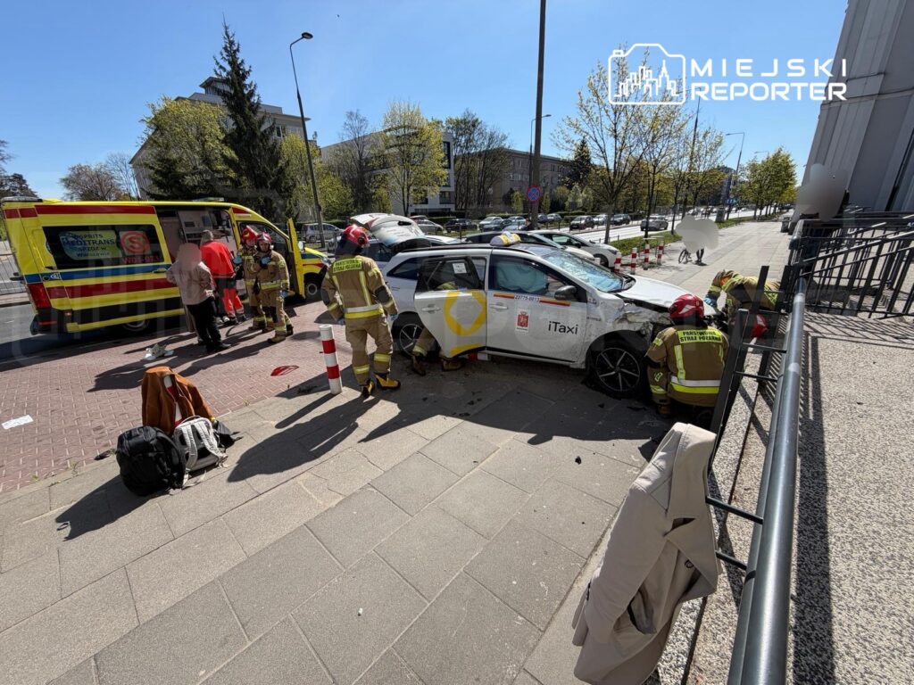 Strażacy udzielają pomocy poszkodowanym w wypadku drogowym obok uszkodzonego taxi na chodniku.