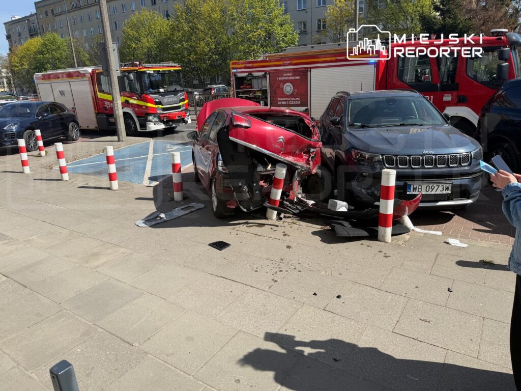Czerwony samochód osobowy zderzony z szarym SUV-em na parkingu, obok strażacki wóz ratunkowy.