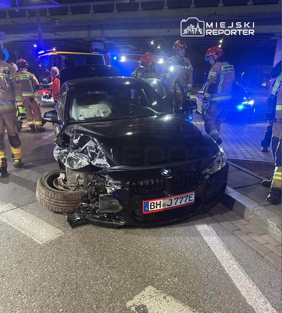 Czarny samochód osobowy z uszkodzoną przednią częścią stoi na drodze, a w tle interweniują strażacy w mundurach.
