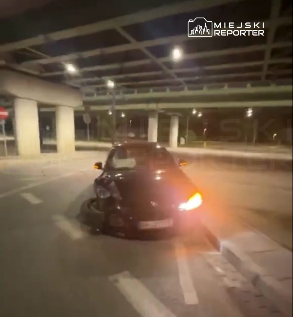 Czarny samochód z uszkodzeniem tyłu stoi na skrzyżowaniu pod wiaduktem, oświetlony lampami ulicznymi.