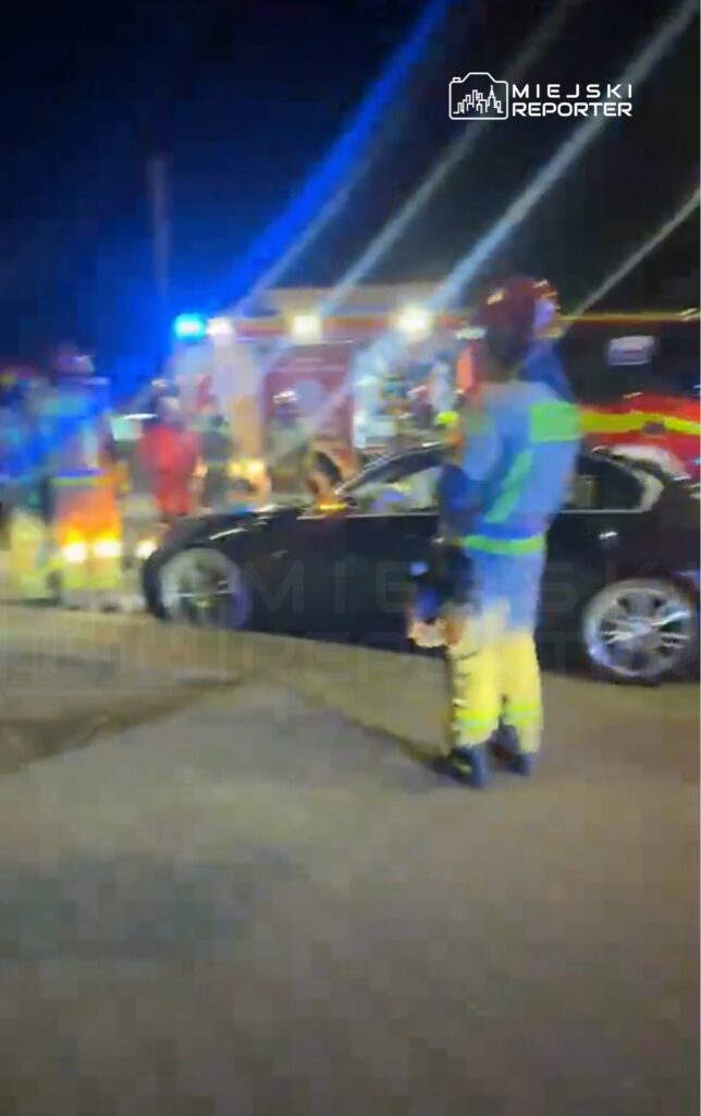 Grupa ratowników w odblaskowych strojach pracuje przy czarnym samochodzie w oświetlonym miejscu nocą.