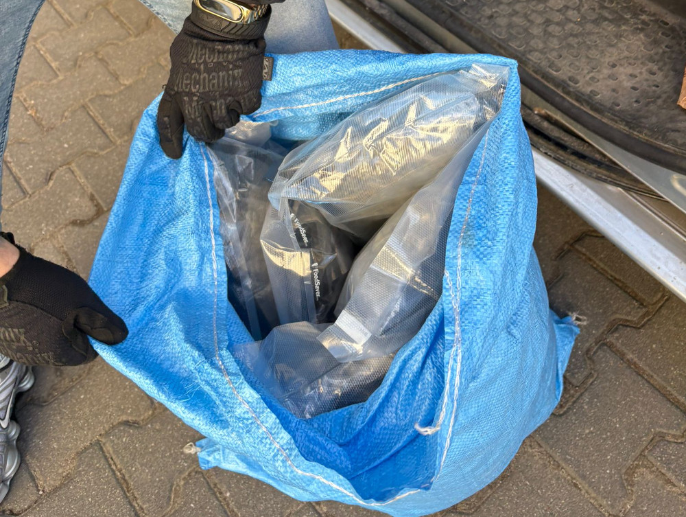 Mężczyzna w rękawicach trzyma niebieski worek, z którego wystają czarne przedmioty, na tle asfaltowej nawierzchni.