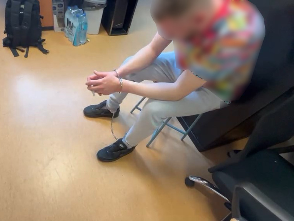 Mężczyzna w kolorowej koszulce siedzi na krześle, z rękami skrępowanymi w kajdankach, na podłodze w pomieszczeniu biurowym.