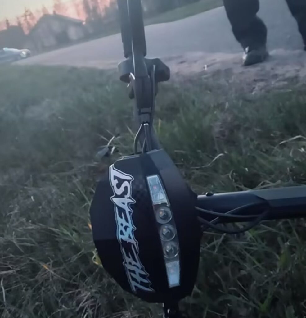 Skuter elektryczny z napisem "THE BEAST" leży na trawie obok drogi, w tle widać samochód i zarys budynków.