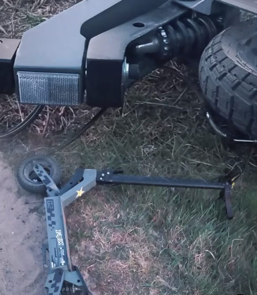 Uszkodzony skuter elektryczny leży na ziemi w trawie, obok pojazdu z dużymi oponami.