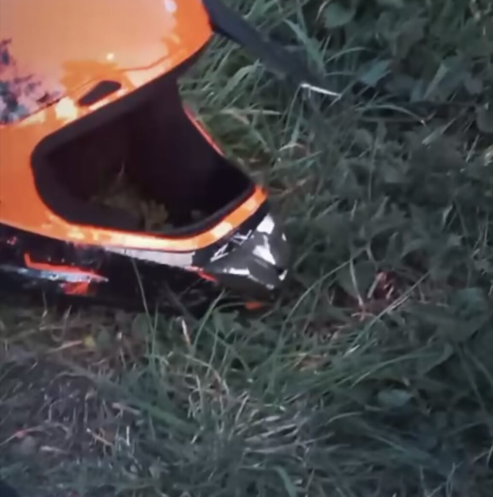 Pomarańczowy kask motocyklowy leży na trawie w otoczeniu zielonych roślin.