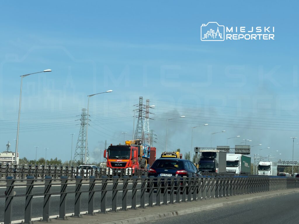 Czerwony ciężarowy pojazd MAN porusza się po ruchliwej autostradzie, obok widocznych innych samochodów i ciężarówek. W tle widać wieże energetyczne oraz dym unoszący się z oddali.
