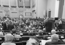 Mężczyzna w garniturze uśmiechający się i trzymający rękę na czole w sali obrad, wśród zgromadzonych ludzi.