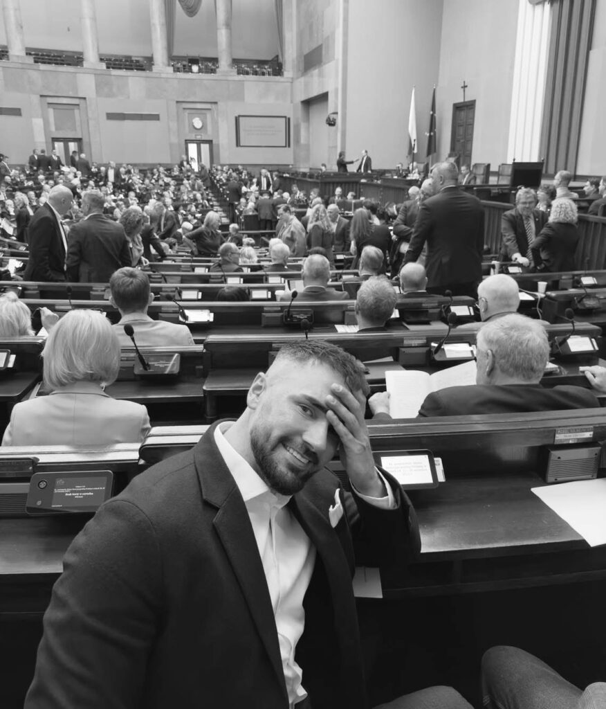 Mężczyzna w garniturze uśmiechający się i trzymający rękę na czole w sali obrad, wśród zgromadzonych ludzi.