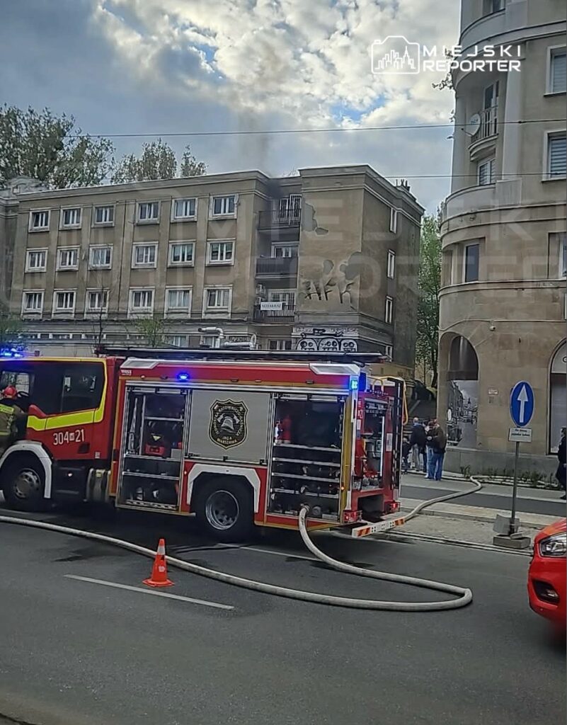 Straż pożarna z włączonymi sygnałami świetlnymi podjeżdża do budynku mieszkalnego, z którego wydobywa się dym.