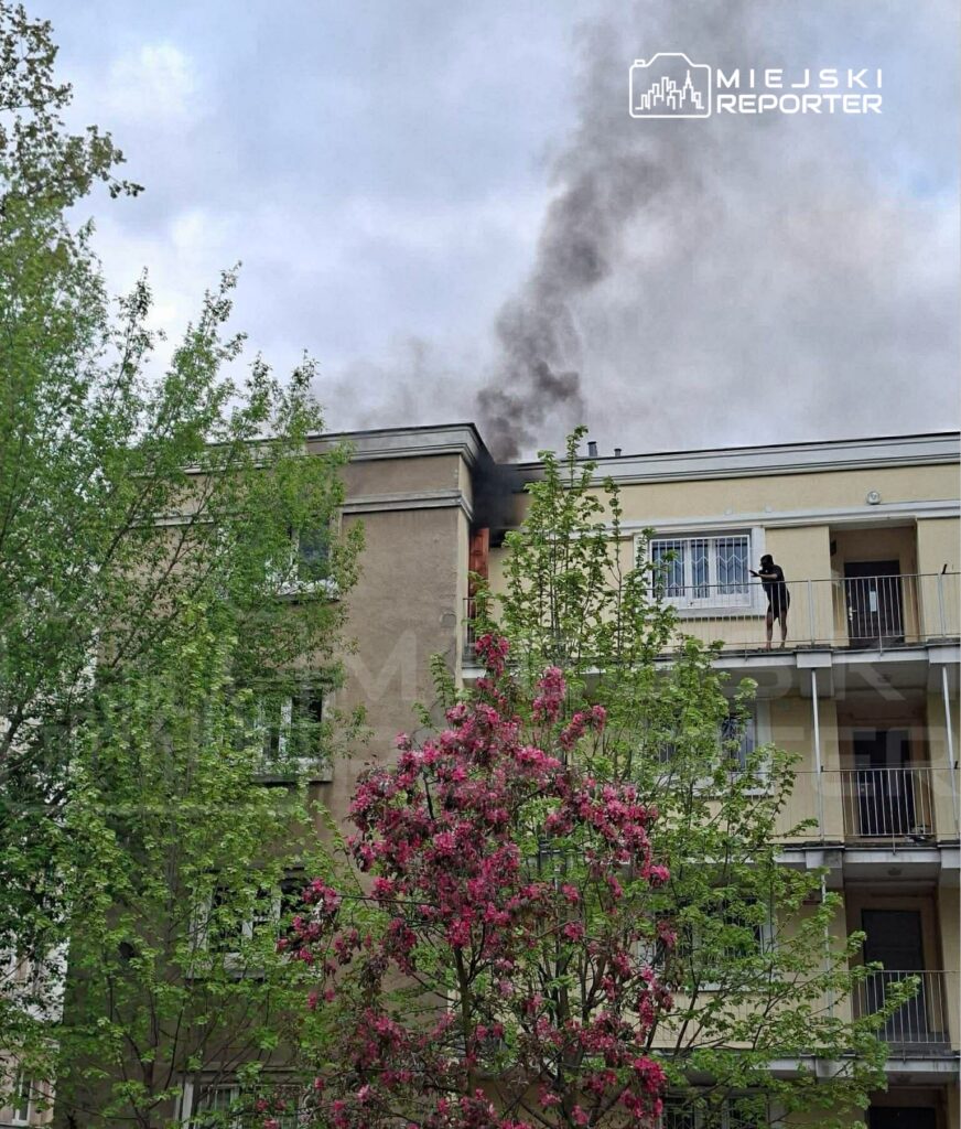 Na balkonie budynku mieszkalnego stoi mężczyzna, a z dachu wydobywa się czarny dym. W tle widać kwitnące drzewa.