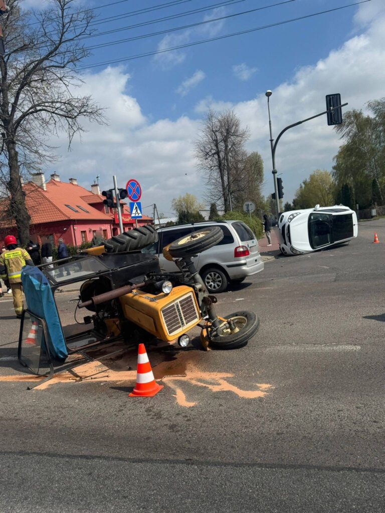 Na skrzyżowaniu leży przewrócony żółty traktor, obok niego widoczny jest biały samochód osobowy. W tle widać czerwony budynek i sygnalizację świetlną. Wokół miejsce zdarzenia zabezpieczają służby ratunkowe.
