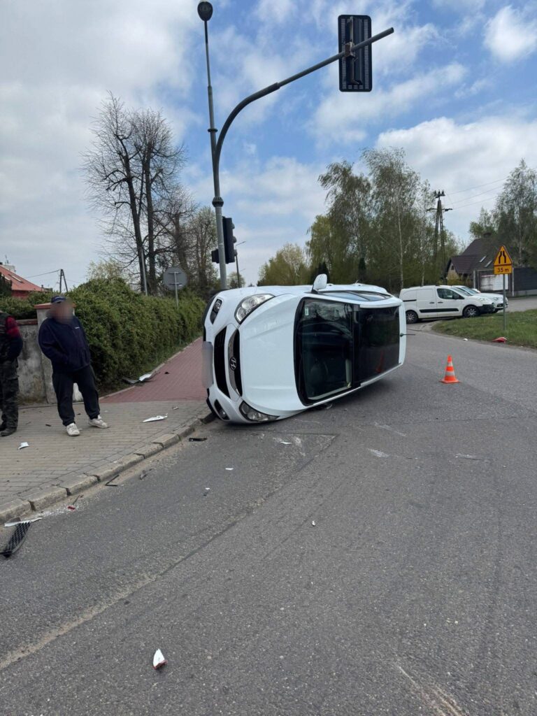 Biały samochód osobowy leży na boku na skrzyżowaniu, obok stoi mężczyzna, a w tle widać znaki drogowe.