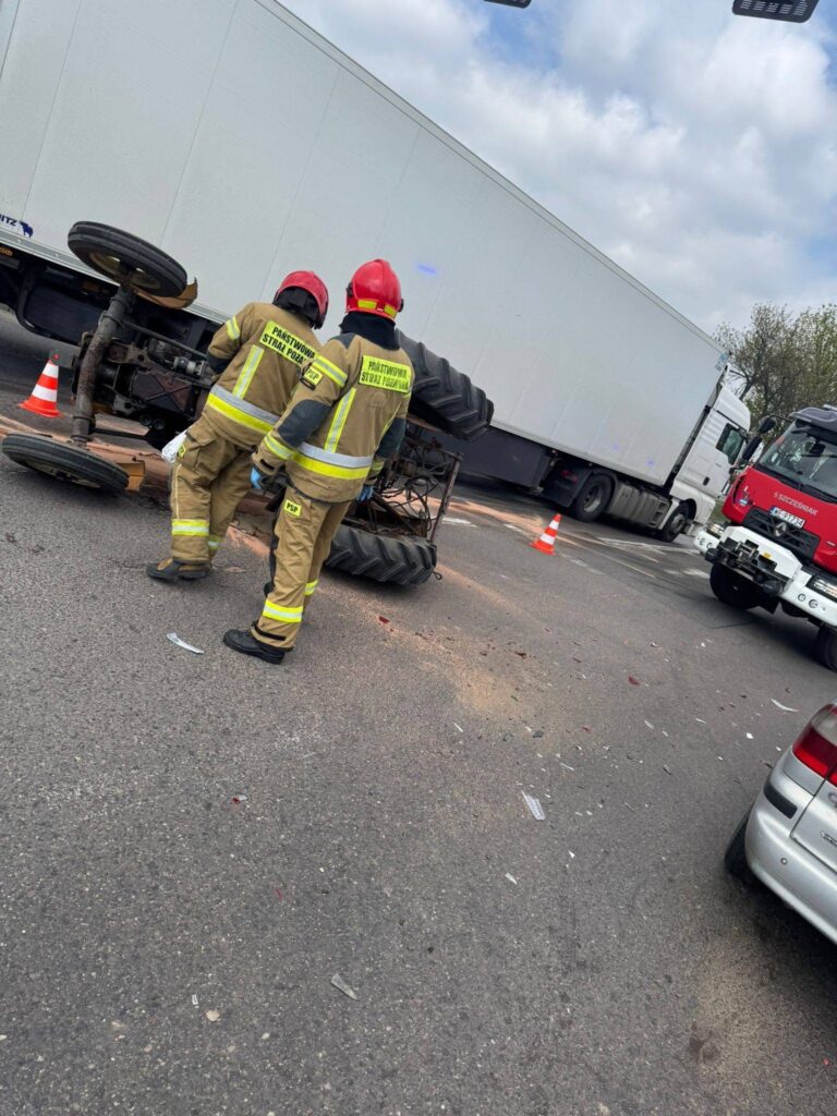 Dwóch strażaków w hełmach bada przewrócone koło ciężarówki na drodze, otoczeni przez rozbite szkło i znaki ostrzegawcze.