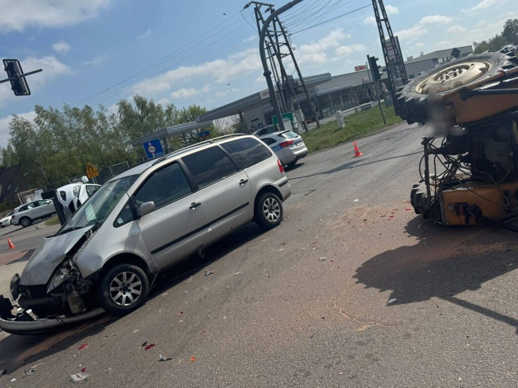 Zniszczony samochód osobowy z uszkodzoną przednią częścią stoi na drodze, obok przewrócony pojazd. W tle widać stację benzynową oraz inne samochody.