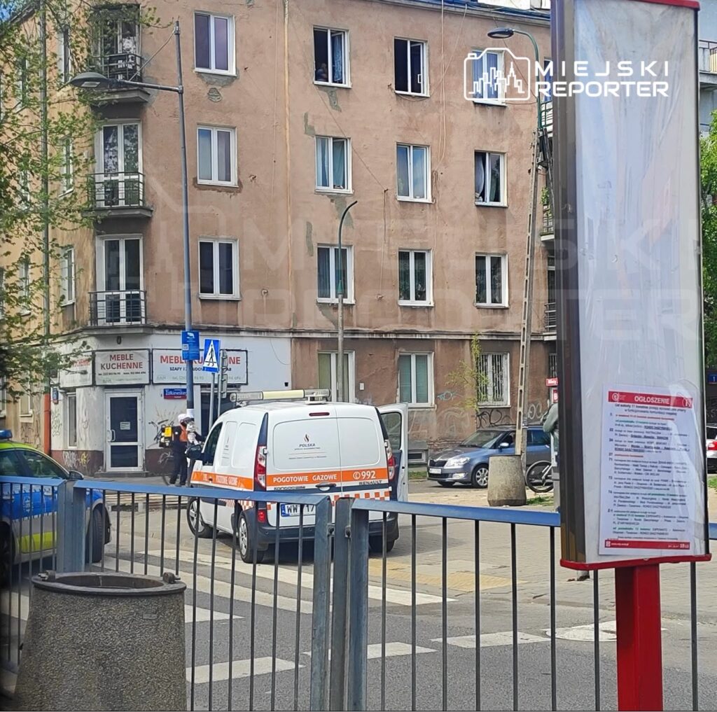 Gazowy zespół ratunkowy bada sytuację przed budynkiem z widocznymi oknami w mieście. W tle stoją różne pojazdy.