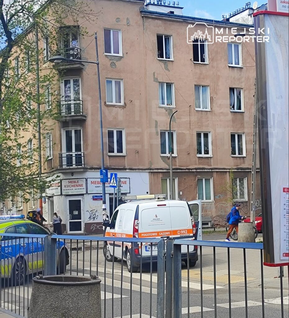 Na ulicy stoi furgonetka pogotowia gazowego, obok policyjny radiowóz, a w oknach budynku widać osoby.