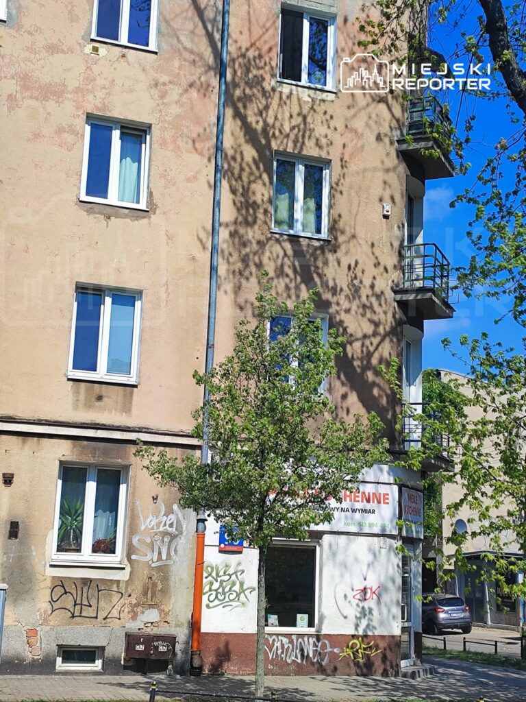 Budynek mieszkalny z graffiti na ścianach, w tle widać drzewa i niebieskie niebo. Na parterze lokal usługowy.