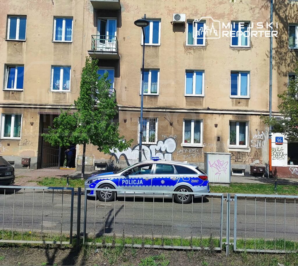 Policjant w mundurze rozmawia z osobą przy wejściu do budynku, obok zaparkowany radiowóz.