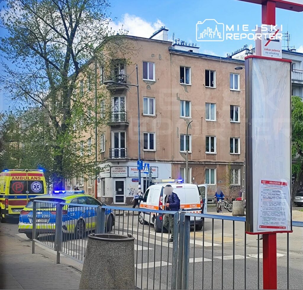 Na ulicy widać pojazdy służb ratunkowych, w tym ambulans i radiowóz policji, obok budynku mieszkalnego.