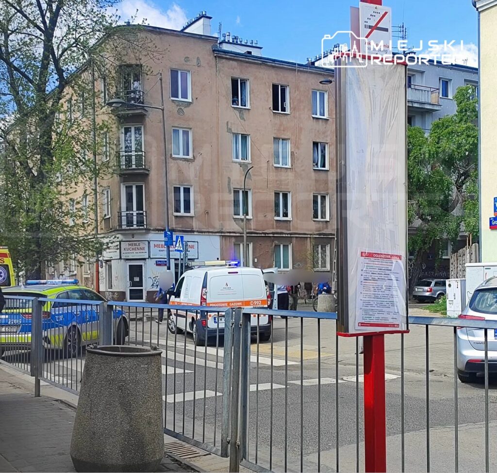 Na ulicy stoi kilka pojazdów policyjnych i ambulansów, a w tle widoczny jest blok mieszkalny z otwartymi oknami.