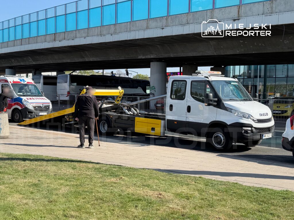 Mężczyzna z laską obserwujący holownik z uszkodzonym pojazdem na parkingu w pobliżu budynku.