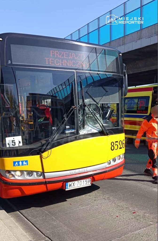 Autobus komunikacji miejskiej z uszkodzoną szybą stoi na ulicy, obok pracownik w pomarańczowym uniformie.