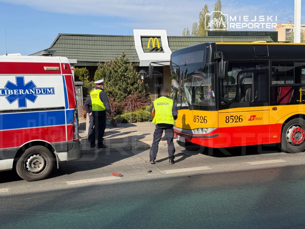 Na skrzyżowaniu stoi autobus z numerem 8526, obok niego znajduje się karetka pogotowia. Policjant w kamizelce „Nadzór Ruchu” rozmawia z innym funkcjonariuszem. W tle widoczna restauracja fast food.