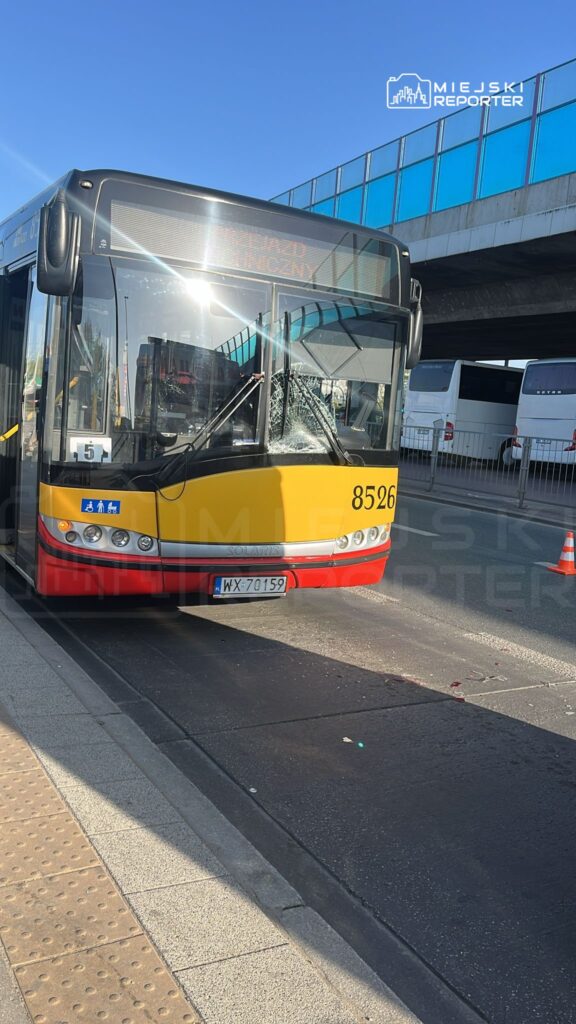 Autobus miejski z numerem 8526 z uszkodzoną szybą stoi na przystanku w słoneczny dzień.