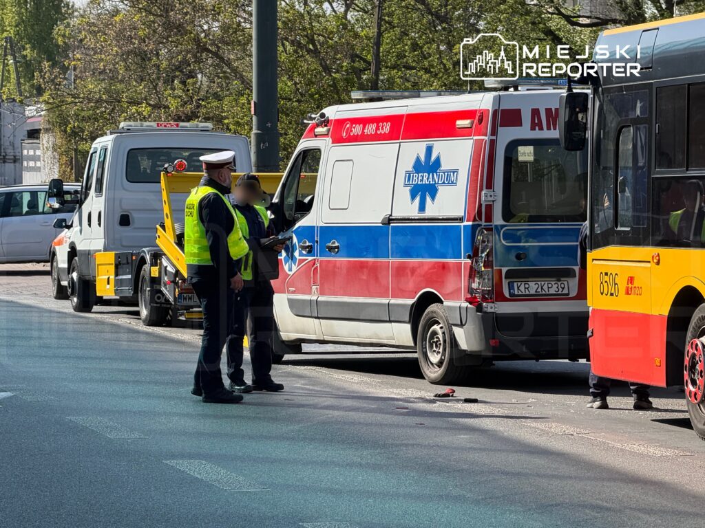 Funkcjonariusz policji rozmawia z ratownikiem medycznym obok karetki, na drodze obok przystanku autobusowego.