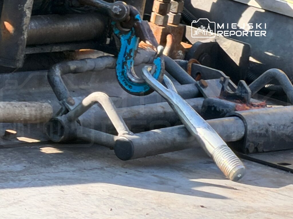 Metalowy hak i pręt mocujący na tle stalowej konstrukcji, używane w pracach budowlanych.