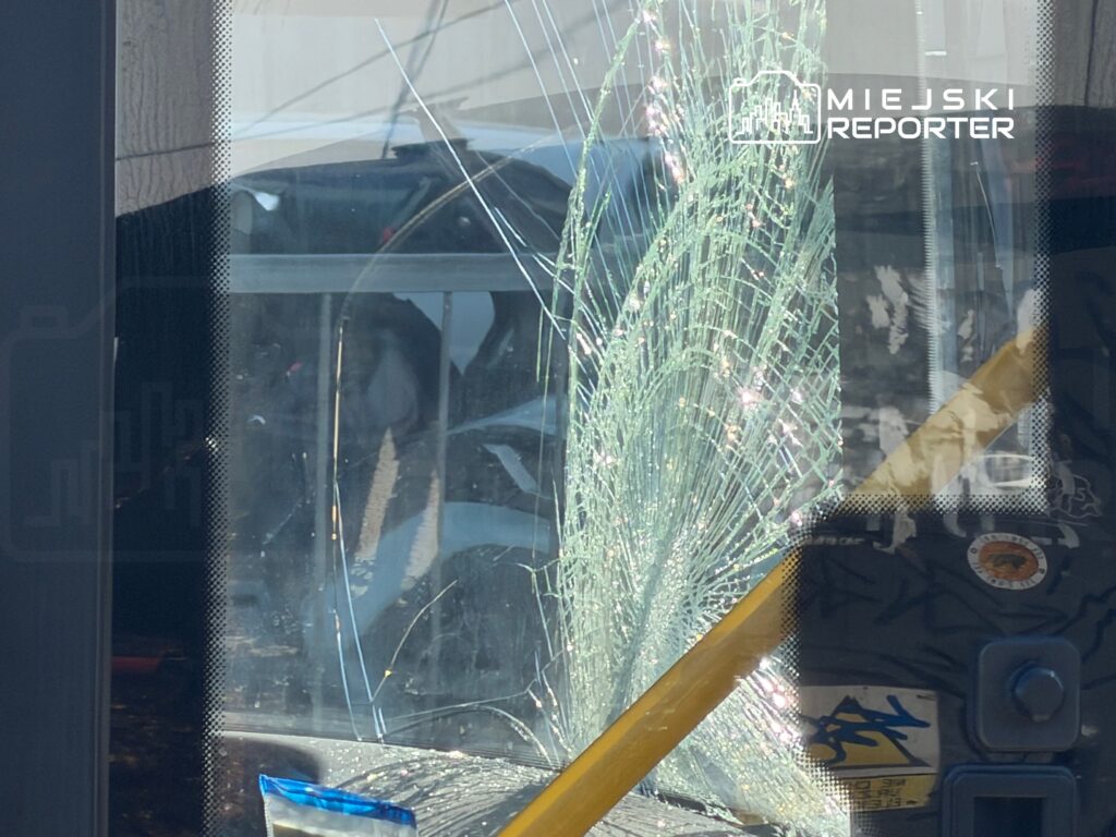 Pęknięta szyba w drzwiach pojazdu, z widocznymi odłamkami szkła i odbiciem otoczenia.