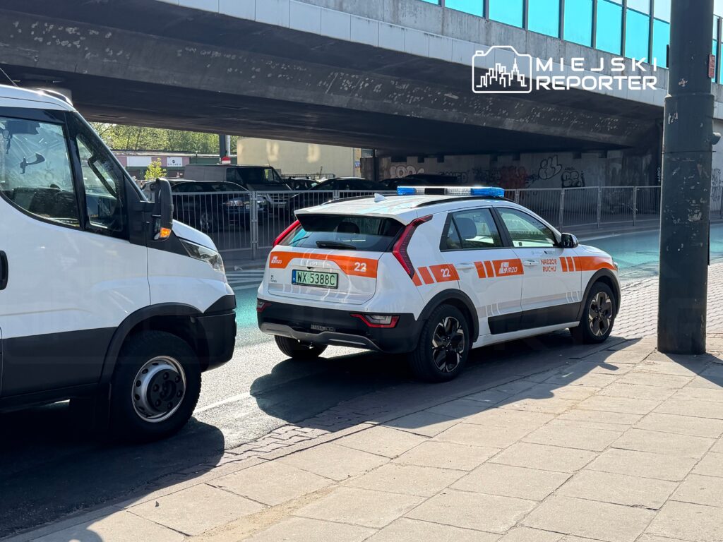 Samochód służbowy z niebieskimi światłami zatrzymany obok białego furgonu pod mostem w mieście.