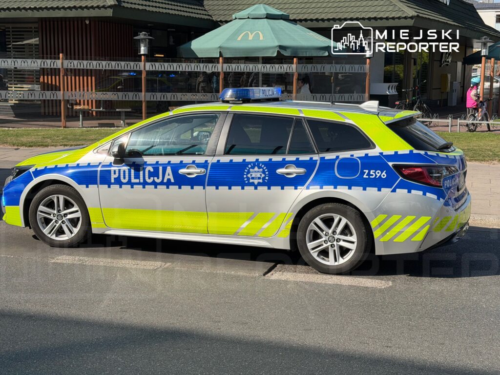 Radiowóz policyjny w biało-niebieskim kolorze stoi na ulicy obok restauracji szybkiej obsługi.
