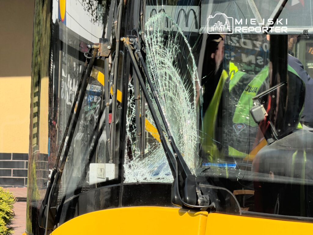 Uszkodzony przód autobusu z pękniętą szybą, w tle widoczni funkcjonariusze policji.