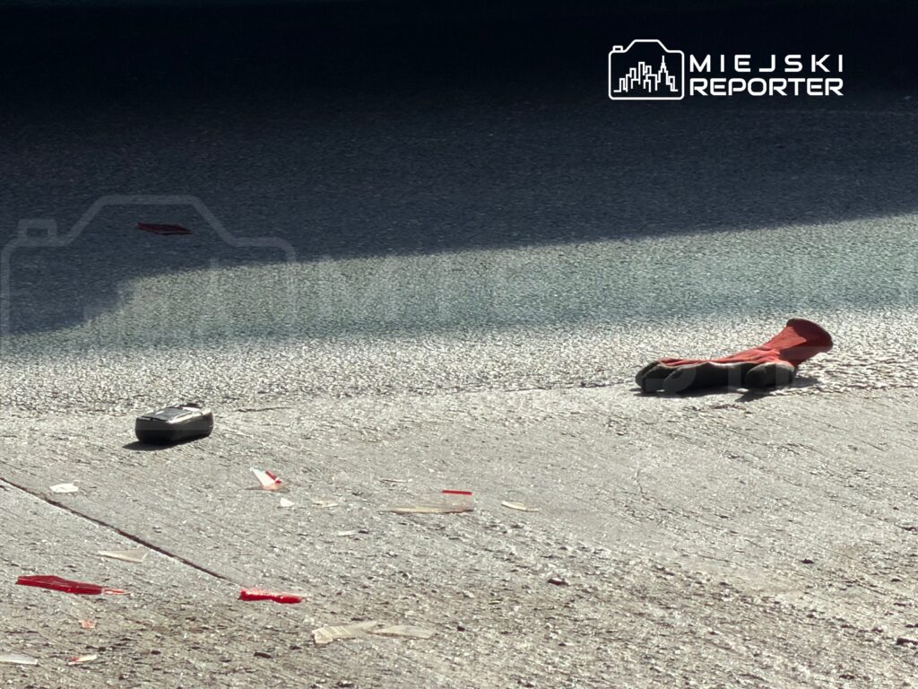 Czerwona rękawica i metalowy przedmiot leżą na szarej nawierzchni, w otoczeniu porozrzucanych kawałków szkła.