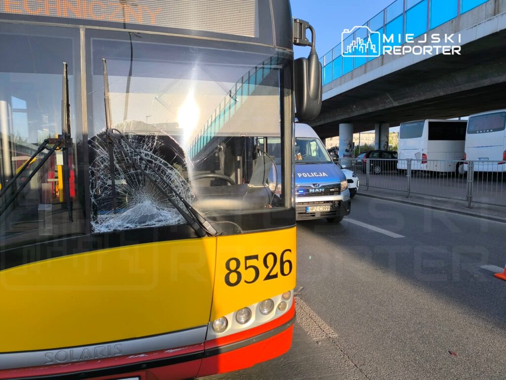 Uszkodzony autobus z pękniętą szybą stoi na ulicy obok radiowozu policyjnego, w tle widoczny most.