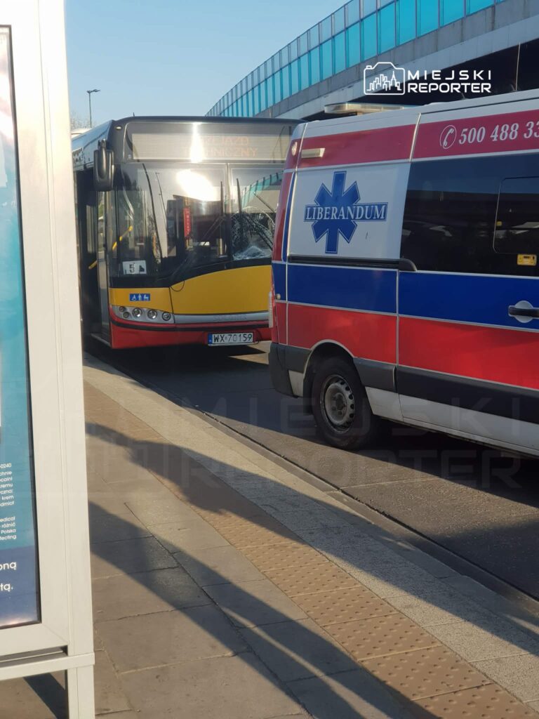Autobus miejski zatrzymany na przystanku obok karetki pogotowia, w tle nowoczesny budynek z przeszkloną fasadą.