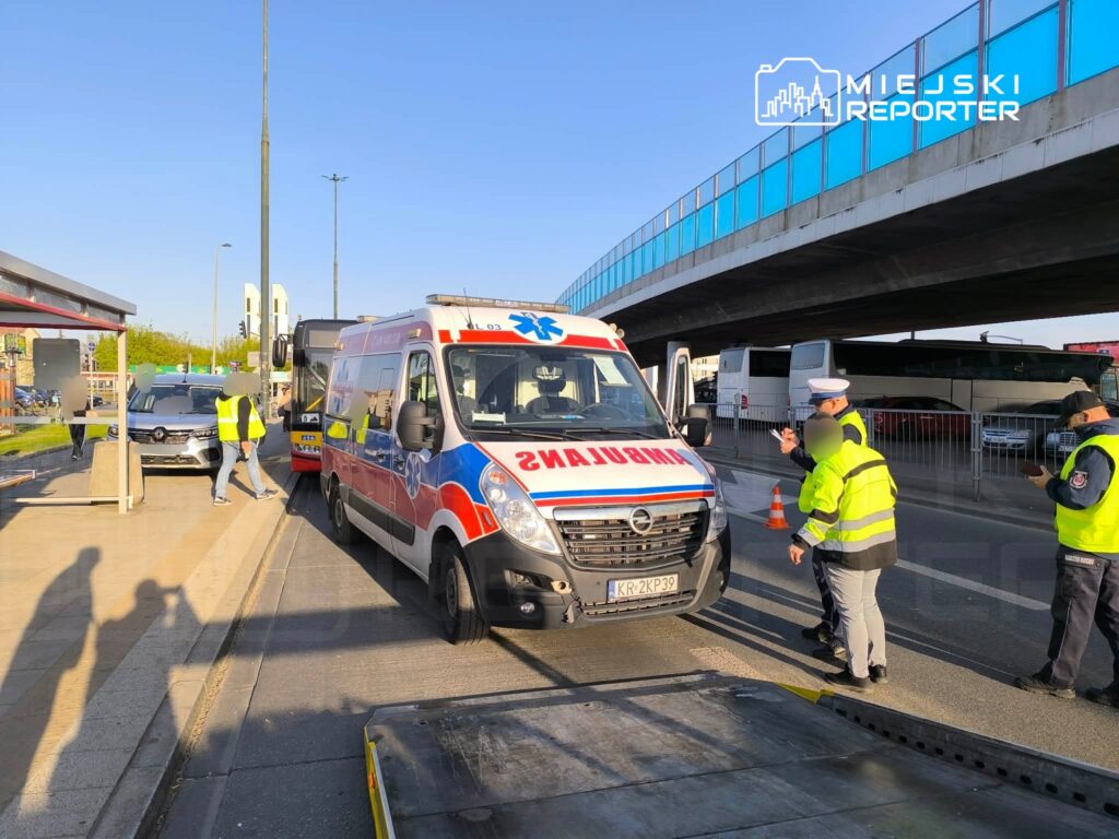 Karetka pogotowia zatrzymana obok przystanku, a pracownicy w żółtych kamizelkach rozmawiają z policjantem.
