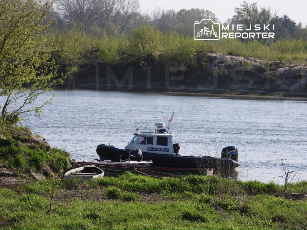Policyjna łódź patrolowa zacumowana przy brzegu rzeki, w otoczeniu zieleni i drzew. W tle widoczna woda.