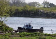 Policyjna łódź patrolowa zacumowana przy brzegu rzeki, w otoczeniu zieleni i drzew. W tle widoczna woda.