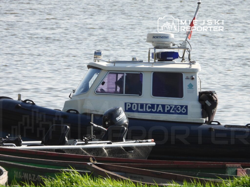 Policyjny kuter motorowy PZ-36 zacumowany przy brzegu rzeki, obok mniejszych łodzi wędkarskich.