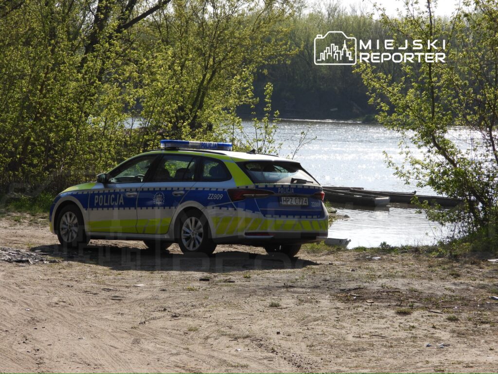 Policyjny radiowóz zaparkowany na brzegu rzeki, otoczony zielonymi drzewami i błyszczącą wodą.