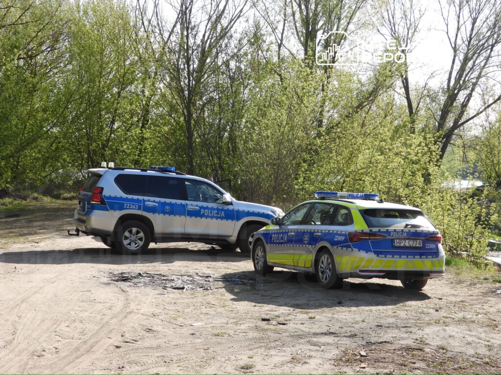 Dwa radiowozy policyjne zaparkowane na piaszczystym terenie otoczonym zielonymi drzewami.