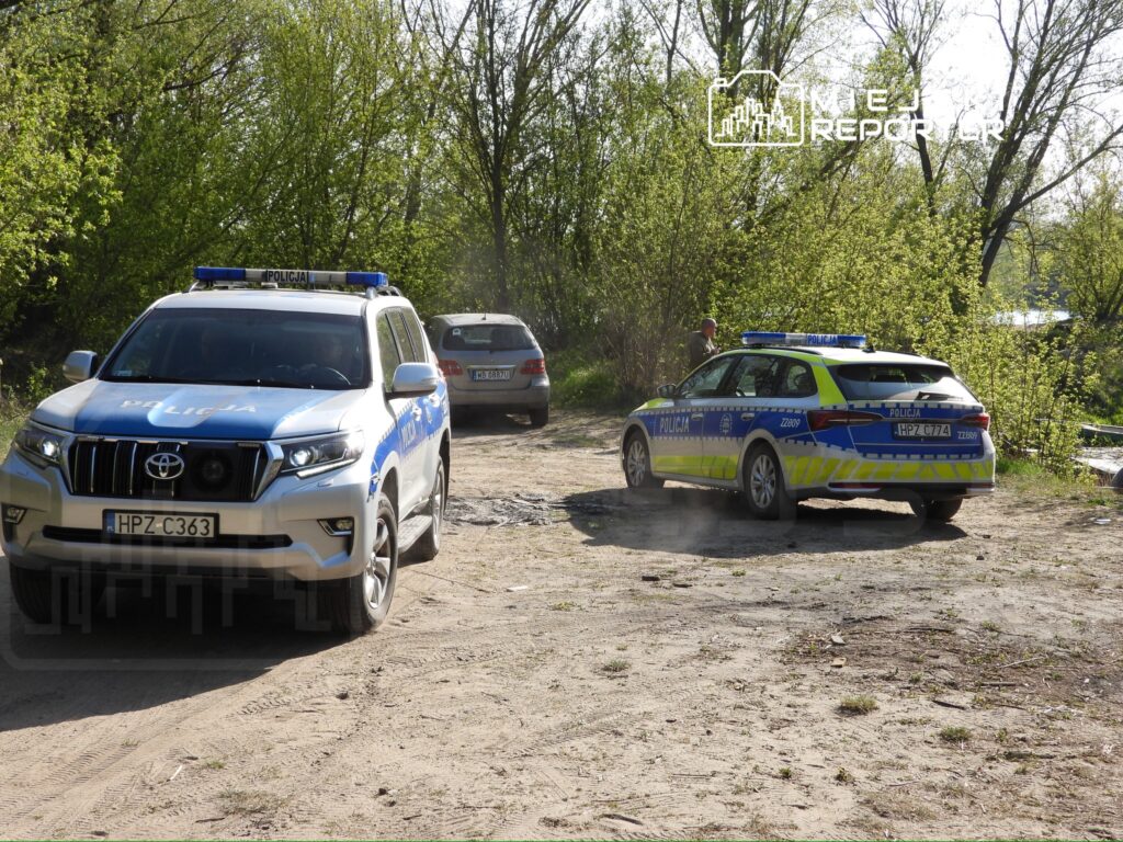 Policyjny SUV oraz radiowóz parkują na piaszczystym terenie otoczonym drzewami, a policjant prowadzi czynności.