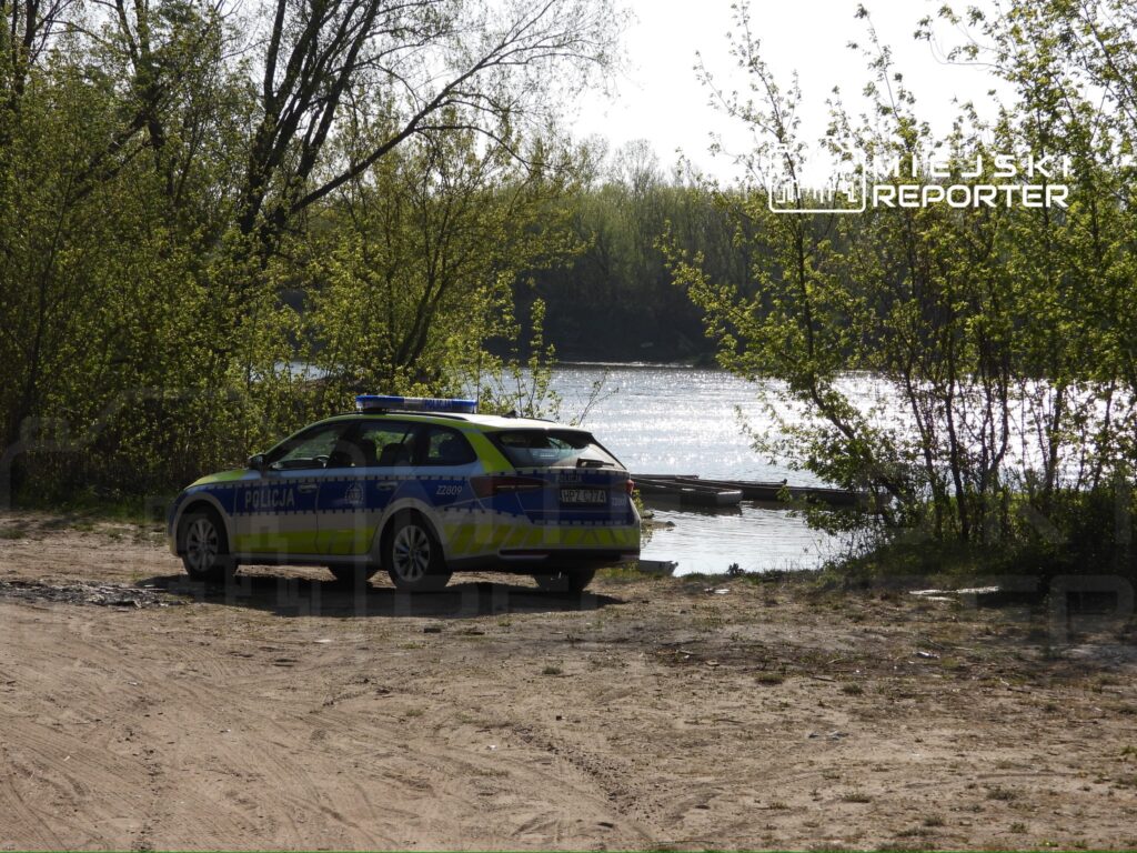 Radiowóz policyjny zaparkowany na piaszczystym brzegu rzeki, w otoczeniu zieleni i drzew.