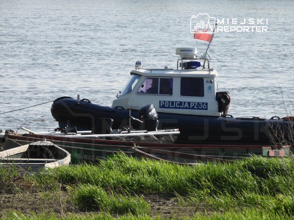 Policyjny kuter motorowy z flagą na wodzie, obok niego mała łódź w otoczeniu zielonej trawy.
